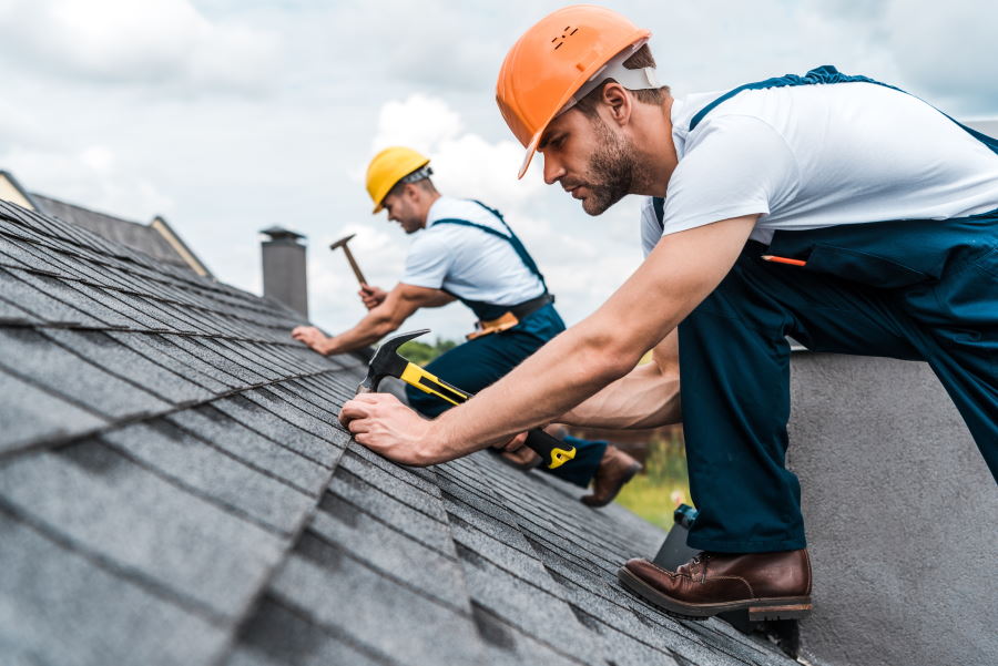 Two roofers installing shingles on a rooftop, one using a hammer and the other securing materials